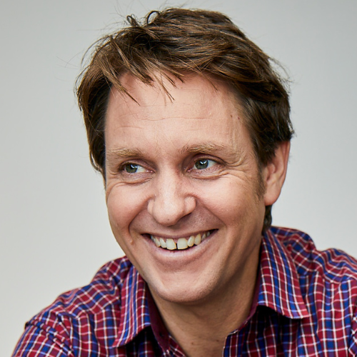 Headshot of Craig Reucassel in a red white, and blue plaid collared shirt. 