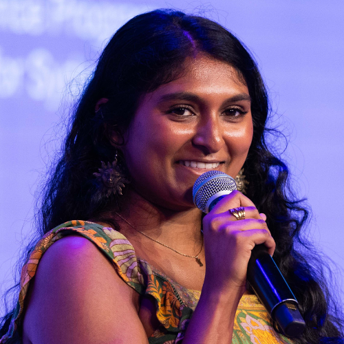Headshot of Grace Vegesana in a floral yellow top holding a microphone. 