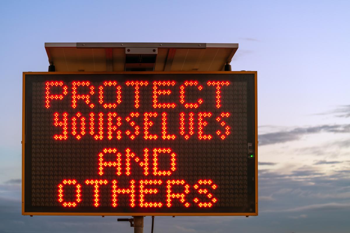 light up road sign that reads 'protect yourselves and others'